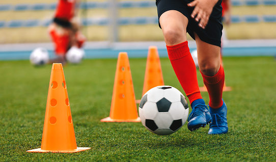 Footballer dribbling ball on training between orange cones. Young football player in sports blue cleats and red socks